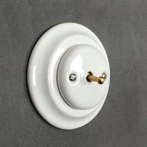 May include: A round white ceramic light switch with a glossy finish, featuring a central brass toggle lever and two small silver-coloured metal screws securing the faceplate. The switch has a stepped circular design with a smooth, reflective surface.