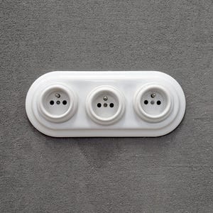 Peut inclure: Une prise électrique blanche avec trois prises. La prise de courant de forme ovale est montée sur un mur gris. Chaque prise comporte trois trous pour les fiches.