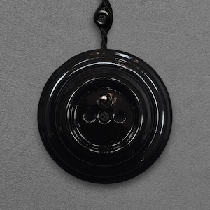 Peut inclure: Un interrupteur de lumière noir et rond avec une finition brillante. L'interrupteur présente un design circulaire avec des anneaux concentriques et trois petits trous. Un cordon et un bouton noirs sont attachés en haut. L'interrupteur est monté sur un mur gris.