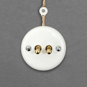 Pode incluir: Um interruptor de luz redondo branco com dois interruptores de alternância de latão e dois parafusos prateados. Uma peça de cerâmica branca a condizer e um cordão de fibra natural são visíveis acima. O fundo é cinzento liso.