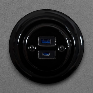Könnte beinhalten: Eine glänzend schwarze USB-Steckdose mit zwei Anschlüssen: einem USB-A und einem USB-C. Die runde Platte hat ein dekoratives Design und ist an einer grauen Wand montiert. Die USB-Anschlüsse haben blaue Innenräume.
