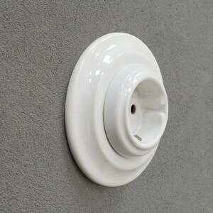 K&ouml;nnte beinhalten: Eine wei&szlig;e Keramiksteckdose mit kreisf&ouml;rmigem Design. Die Steckdose hat eine gl&auml;nzende Oberfl&auml;che und ist an einer grau strukturierten Wand montiert. Die Steckdose hat ein zentrales Loch f&uuml;r den Stecker.