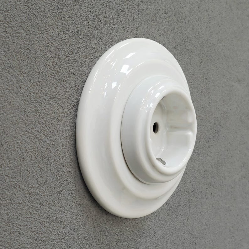 K&ouml;nnte beinhalten: Eine wei&szlig;e Keramiksteckdose mit kreisf&ouml;rmigem Design. Die Steckdose hat eine gl&auml;nzende Oberfl&auml;che und ist an einer grau strukturierten Wand montiert. Die Steckdose hat ein zentrales Loch f&uuml;r den Stecker.