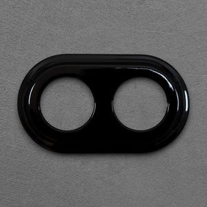 Könnte beinhalten: Ein glänzend schwarzes, ovales Objekt mit zwei kreisförmigen Öffnungen. Das Objekt hat einen erhöhten, abgerundeten Rand und befindet sich vor einem grauen Hintergrund. Das Design ist schlicht und modern.