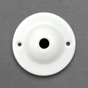Pode incluir: Uma placa de parede circular de cerâmica branca com um acabamento liso e brilhante e um centro elevado em forma de cúpula. A peça inclui uma única abertura circular centralizada e dois orifícios simétricos menores nas laterais para montagem.