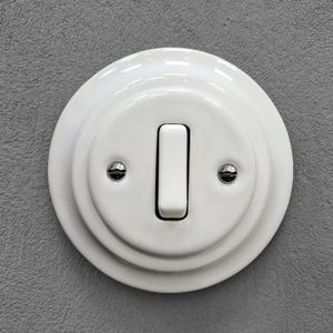 Op de afbeelding: Een ronde witte keramische lichtschakelaar gemonteerd op een muur. De schakelaar is voorzien van een centrale verticale tuimelschakelaar en twee kleine zilverkleurige metalen schroeven aan weerszijden om de afdekplaat vast te zetten.