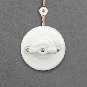 May include: A white ceramic light switch with a cross-shaped handle and a matching pull cord. The switch is mounted on a gray wall. The design is a classic, vintage style.