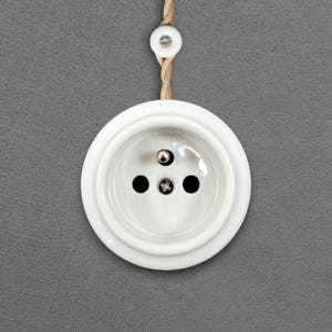Peut inclure: Une prise électrique en céramique blanche avec trois trous et un contact métallique central. La prise est circulaire avec un bord surélevé, suspendue par un cordon beige torsadé. Un bouton en céramique assorti est en haut du cordon.