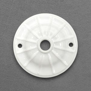 Puede incluir: Una roseta de techo blanca y circular con un agujero central y dos agujeros más pequeños. La roseta presenta un diseño acanalado que irradia desde el centro, creando una superficie texturizada. El objeto es de cerámica o porcelana.