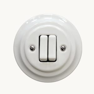 May include: A circular white ceramic double light switch featuring two vertical toggle switches in the centre. The switch plate is secured with two silver-coloured metal screws positioned on the left and right sides of the toggle mechanism.