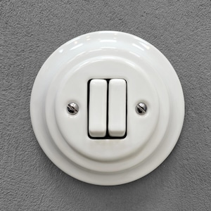 Op de afbeelding: Een witte, ronde dubbele lichtschakelaar met een glanzende afwerking. De schakelaar heeft twee verticale tuimelschakelaars die naast elkaar in het midden zijn geplaatst, vastgezet met twee kleine metalen schroeven met platte kop.
