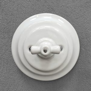 May include: A circular white ceramic light switch with a central rotating toggle handle. The switch features a stepped, tiered base design and is secured with two visible metallic screws on either side of the central horizontal handle mechanism.