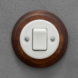 Op de afbeelding: Een lichtschakelaar in vintage-stijl met een witte tuimelschakelaar en een witte, ovale frontplaat. De frontplaat is geplaatst in een ronde, donker gebeitste houten basis. Twee kleine schroeven bevestigen de frontplaat aan de houten basis.
