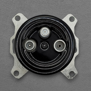 Könnte beinhalten: Ein schwarzes, kreisförmiges Bauteil mit drei Koaxialanschlüssen und einer zentralen Schraube. Es ist auf einer silberfarbenen, vierseitigen Halterung mit abgerundeten Ecken und Befestigungslöchern montiert. Das Bauteil hat eine glänzende Oberfläche.