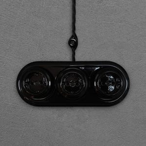 Peut inclure: Une plaque de prise électrique noire, de forme ovale, avec trois prises circulaires. La plaque est brillante et fixée sur un mur gris. Un fil noir tressé s'étend du haut, relié à un petit dispositif rond.