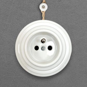 Peut inclure: Une prise électrique blanche et ronde avec un design à trois trous, montée sur un mur gris. La prise a un design décoratif en couches et est suspendue par une corde en fibre naturelle.
