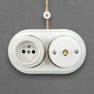 Può includere: Una placca a muro doppia in ceramica bianca dotata di una presa elettrica e un interruttore a levetta. L'unità è montata su una superficie grigia e include un cavo in tessuto intrecciato color corda che sale verso l'alto con un fermacavo in ceramica.