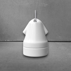 May include: A white ceramic pendant light fixture with a distinctive design. The fixture features a rounded base and a top shaped like a small shirt, with a metal loop for hanging. The overall aesthetic is minimalist and modern.