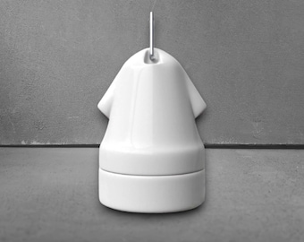May include: A white ceramic bell-shaped object with a smooth, glossy finish. The piece features a rounded top with a small vertical metal loop attached at the apex and a cylindrical base section that appears to be a separate, detachable component.