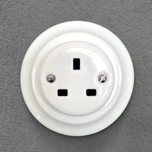 Könnte beinhalten: Eine weiße Steckdose mit drei rechteckigen Löchern und einem kreisförmigen Design. Die Steckdose ist an einer grauen Wand montiert. Zwei Schrauben befestigen die Steckdose an der Wand.