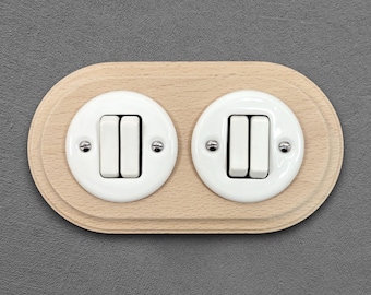 May include: A double light switch assembly featuring two white circular ceramic plates mounted on a single oval light-coloured wooden base. Each plate contains two vertical white toggle switches secured by two small silver-toned metal screws.