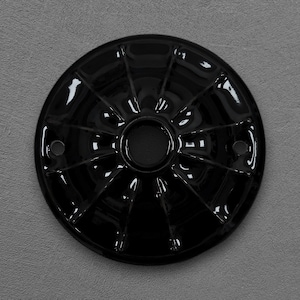 Pode incluir: Uma placa metálica circular preta brilhante com uma abertura central e dois orifícios circulares menores em lados opostos. A superfície apresenta uma série de nervuras radiais em relevo que se estendem do centro até a borda externa.