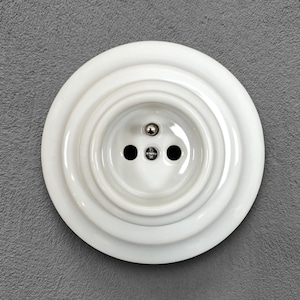 Può includere: Una presa elettrica in ceramica bianca con tre fori circolari e un perno di messa a terra centrale. La presa è posta su uno sfondo grigio. Il design presenta anelli concentrici, creando un effetto a strati.
