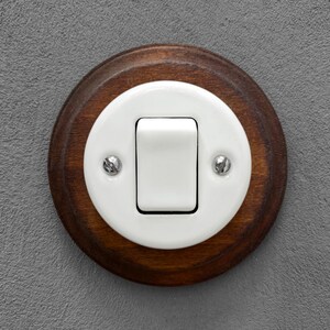May include: A vintage-style light switch with a white toggle and a white ceramic plate, set within a round, dark-stained wooden base. Two silver screws secure the plate to the wood. The switch is mounted on a gray wall.