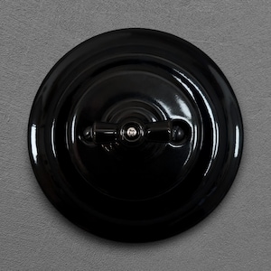 Può includere: Un interruttore della luce nero lucido e rotondo con una leva a bilanciere. L'interruttore è montato su un muro grigio. L'interruttore ha un design classico e vintage.