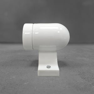 Op de afbeelding: Een witte, cilindrische wandlamp met een gladde, glanzende afwerking. De armatuur heeft een afgerond uiteinde en een platte voetplaat met een enkele zichtbare metalen schroef in het midden aan de onderkant voor bevestiging aan een muur.