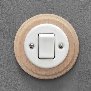 Könnte beinhalten: Ein weißer Keramik-Lichtschalter mit rechteckigem Kippschalter, montiert auf einer runden, hellen Holzbasis. Der Schalter ist mit zwei silbernen Schrauben befestigt. Schlichtes und klassisches Design, passend für verschiedene Einrichtungsstile.