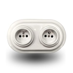 May include: A white double electrical wall socket with a smooth, glossy finish. The unit features two circular recessed sockets, each containing two pin holes and a central grounding pin, set within an oval-shaped, moulded plastic faceplate.