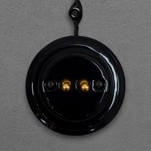 May include: A black, round light switch with three gold-colored buttons and two black buttons. The switch is mounted on a gray wall and has a black cord attached to the top.
