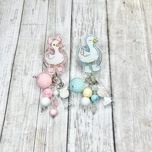 Puede incluir: Dos carretes de insignia acr&iacute;licos con dise&ntilde;os de patos. Uno lleva un lazo y botas rosas, el otro botas azules. Cada carrete tiene un dije de cuentas en colores pastel, incluyendo rosa, blanco y azul.