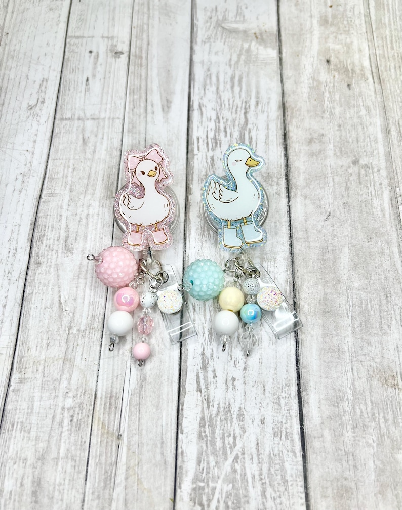 Puede incluir: Dos carretes de insignia acr&iacute;licos con dise&ntilde;os de patos. Uno lleva un lazo y botas rosas, el otro botas azules. Cada carrete tiene un dije de cuentas en colores pastel, incluyendo rosa, blanco y azul.