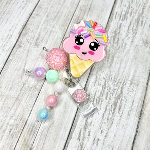 Op de afbeelding: Een roze ijsje badge reel met een schattig gezicht en kleurrijke sprinkles. De badge reel heeft een doorzichtige plastic houder en een kralen bedel met roze, paarse en blauwe kralen. De achtergrond is een lichte houtnerf.
