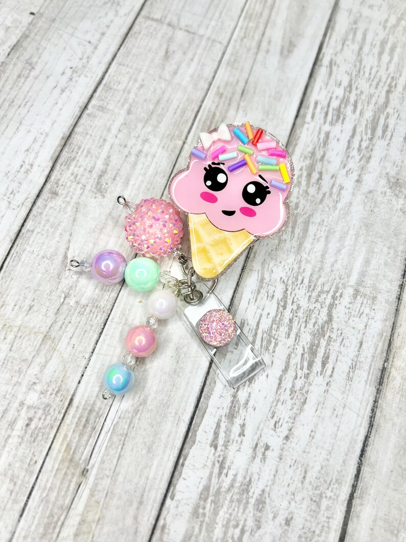 Op de afbeelding: Een roze ijsje badge reel met een schattig gezicht en kleurrijke sprinkles. De badge reel heeft een doorzichtige plastic houder en een kralen bedel met roze, paarse en blauwe kralen. De achtergrond is een lichte houtnerf.