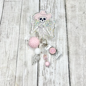 Peut inclure: Un squelette en paillettes rose et blanc portant un chapeau de cowboy rose avec un porte-badge en paillettes rose et blanc. Le porte-badge a un support en plastique transparent et un charme en paillettes rose.