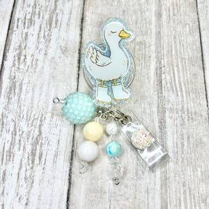 Puede incluir: Un portatarjetas con un dise&ntilde;o de pato azul claro con un contorno brillante y botas a juego. El portatarjetas tiene un clip de pl&aacute;stico transparente y est&aacute; adornado con cuentas en tonos azules, blancos y amarillos.