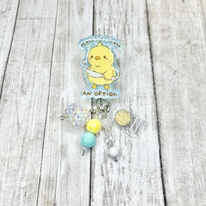 May include: A badge reel with a yellow chick holding a knife, with the text "Peace was never an option." The reel has a beaded charm with iridescent, yellow, blue, and white beads. The background is a light wood grain.