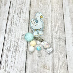 Puede incluir: Un portatarjetas con un dise&ntilde;o de pato blanco sobre un fondo azul claro y purpurina. El pato lleva botas azules y tiene un pico amarillo. El portatarjetas tiene cuentas pastel y un clip de pl&aacute;stico transparente.