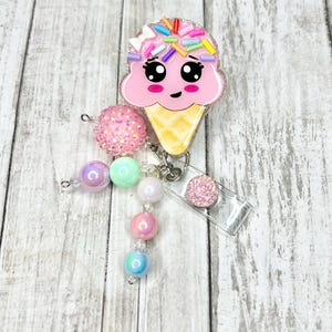 Op de afbeelding: Een roze ijsje badge reel met een lachend gezicht en kleurrijke hagelslag. De reel is bevestigd aan een kralen bedel met roze, groene en iriserende kralen. Een kleine, glinsterende roze cirkel is ook bevestigd.