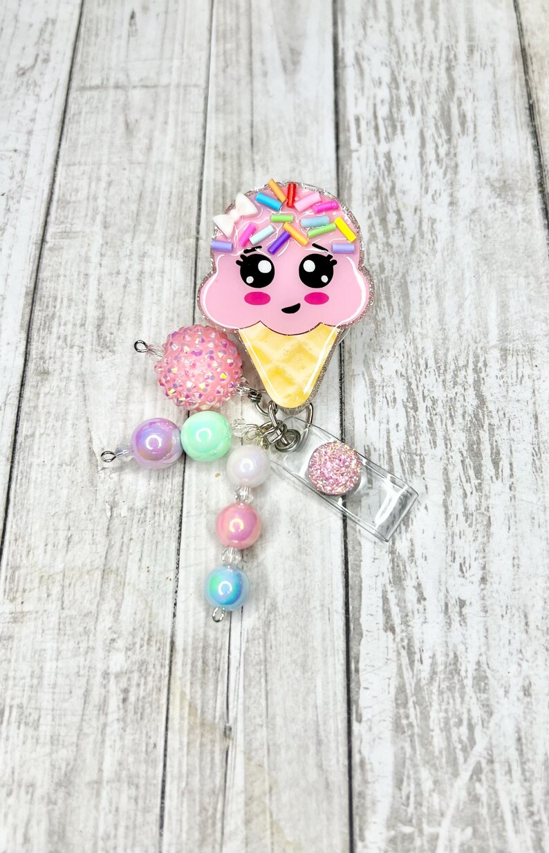 Op de afbeelding: Een roze ijsje badge reel met een lachend gezicht en kleurrijke hagelslag. De reel is bevestigd aan een kralen bedel met roze, groene en iriserende kralen. Een kleine, glinsterende roze cirkel is ook bevestigd.