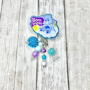 May include: A blue and purple badge reel featuring a cartoon bear on a cloud with the text "Born Grumpy." The badge reel has a variety of dangling beads in shades of blue, purple, and white.
