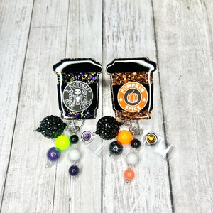May include: Two Halloween-themed badge reels shaped like coffee cups. One features a "Nightmare Before Coffee" design, the other says "Pumpkin Spice". Each has a beaded charm with black, purple, orange, and white beads.