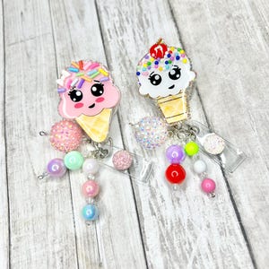 Op de afbeelding: Twee badge-rolhouders in de vorm van een ijshoorntje met lachende gezichten. E&eacute;n is roze met hagelslag, de andere wit met een kers erop. Elk heeft een kralen dangle in pastelkleuren en een doorzichtige plastic rol.