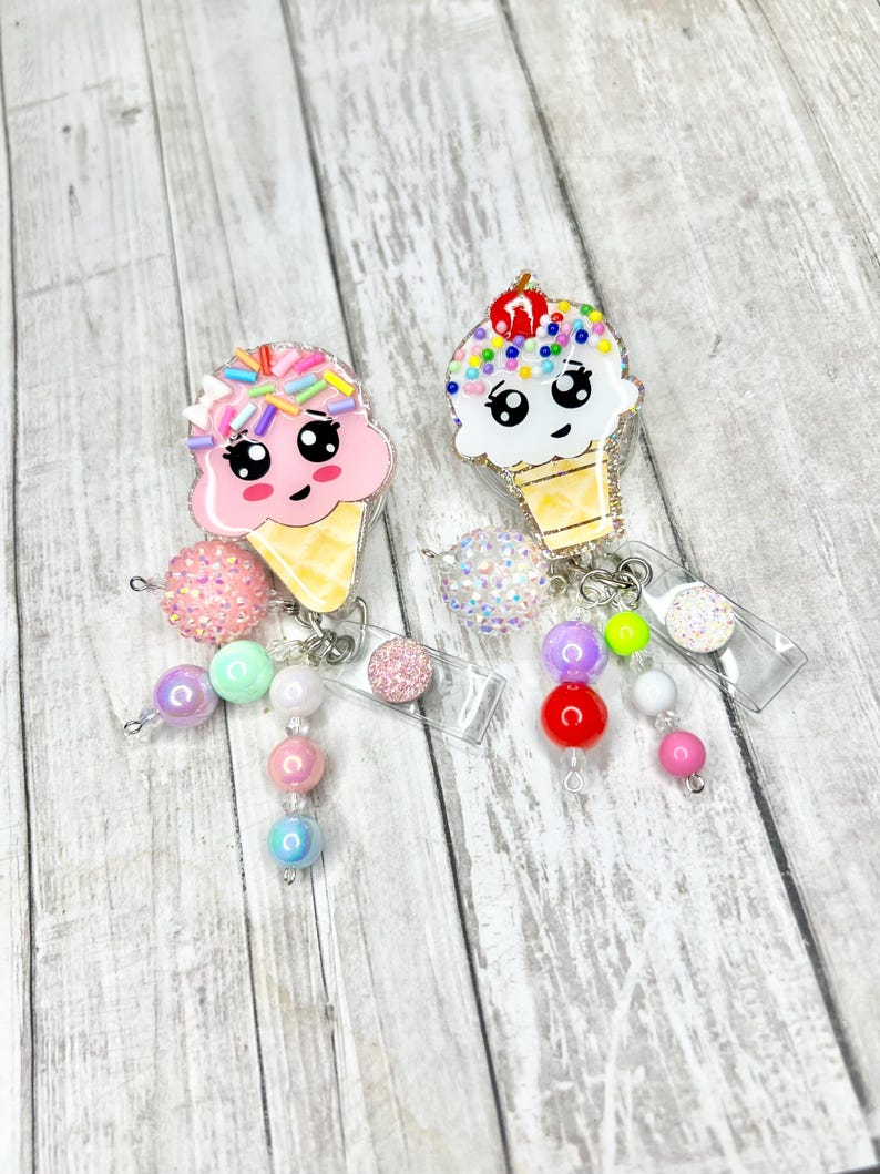 Op de afbeelding: Twee badge-rolhouders in de vorm van een ijshoorntje met lachende gezichten. E&eacute;n is roze met hagelslag, de andere wit met een kers erop. Elk heeft een kralen dangle in pastelkleuren en een doorzichtige plastic rol.