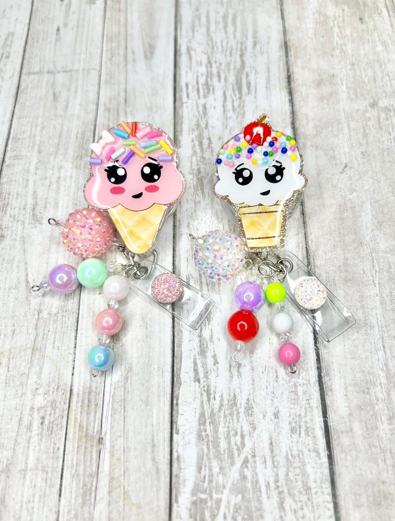 Op de afbeelding: Twee badgehaspels in de vorm van een ijshoorntje met lachende gezichten. De ene heeft roze ijs met hagelslag, de andere heeft wit ijs met kleurrijke hagelslag en een kers. Elk heeft een kralen dangle met verschillende kleuren.