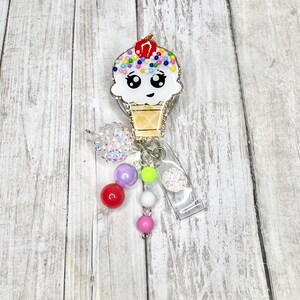Op de afbeelding: Een schattige badge reel in de vorm van een ijshoorntje met een lachend gezicht, kleurrijke hagelslag en een kers. De badge reel heeft een doorzichtige plastic clip en een kralenbedel.
