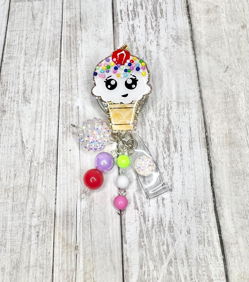 Op de afbeelding: Een schattige badge reel in de vorm van een ijshoorntje met een lachend gezicht, kleurrijke hagelslag en een kers. De badge reel heeft een doorzichtige plastic clip en een kralenbedel.
